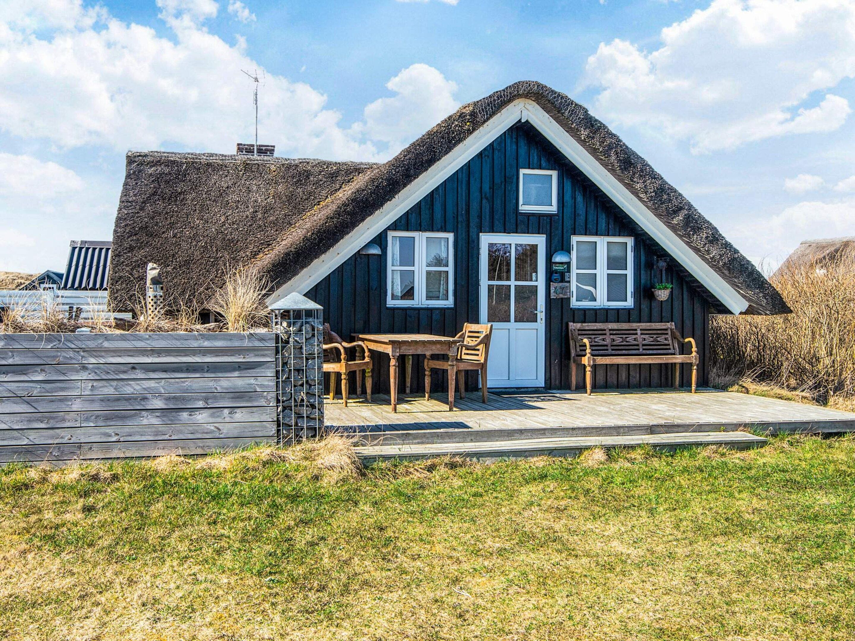 A wooden house with a thatched roof, a wooden bench, and a table with chairs outside.