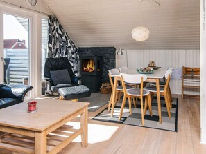 A cozy living room with a fireplace, a dining area with wooden chairs, and a sofa with a black and white patterned throw. - Modish Holiday Home in Jutland near Sea (Harboore)