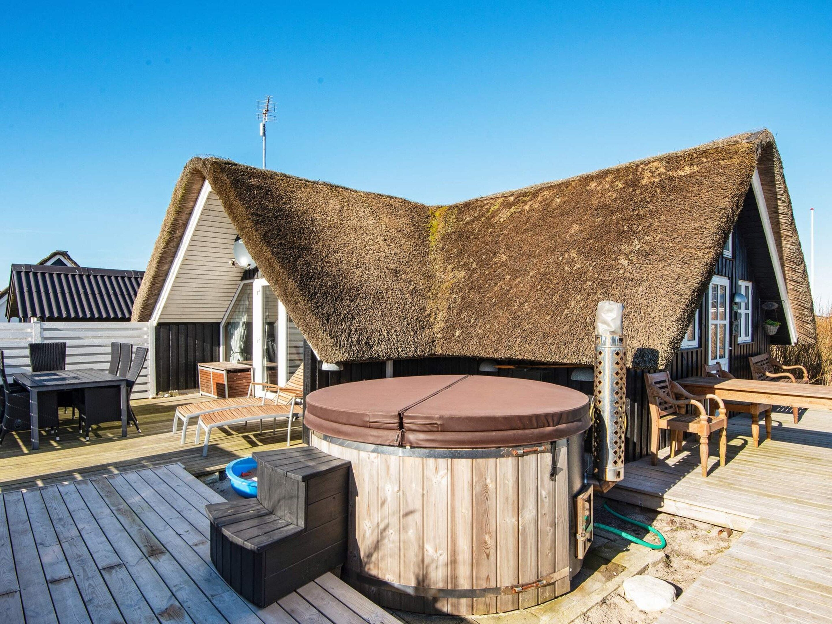 Una terraza de madera con jacuzzi, asientos al aire libre y una estructura con techo de paja.