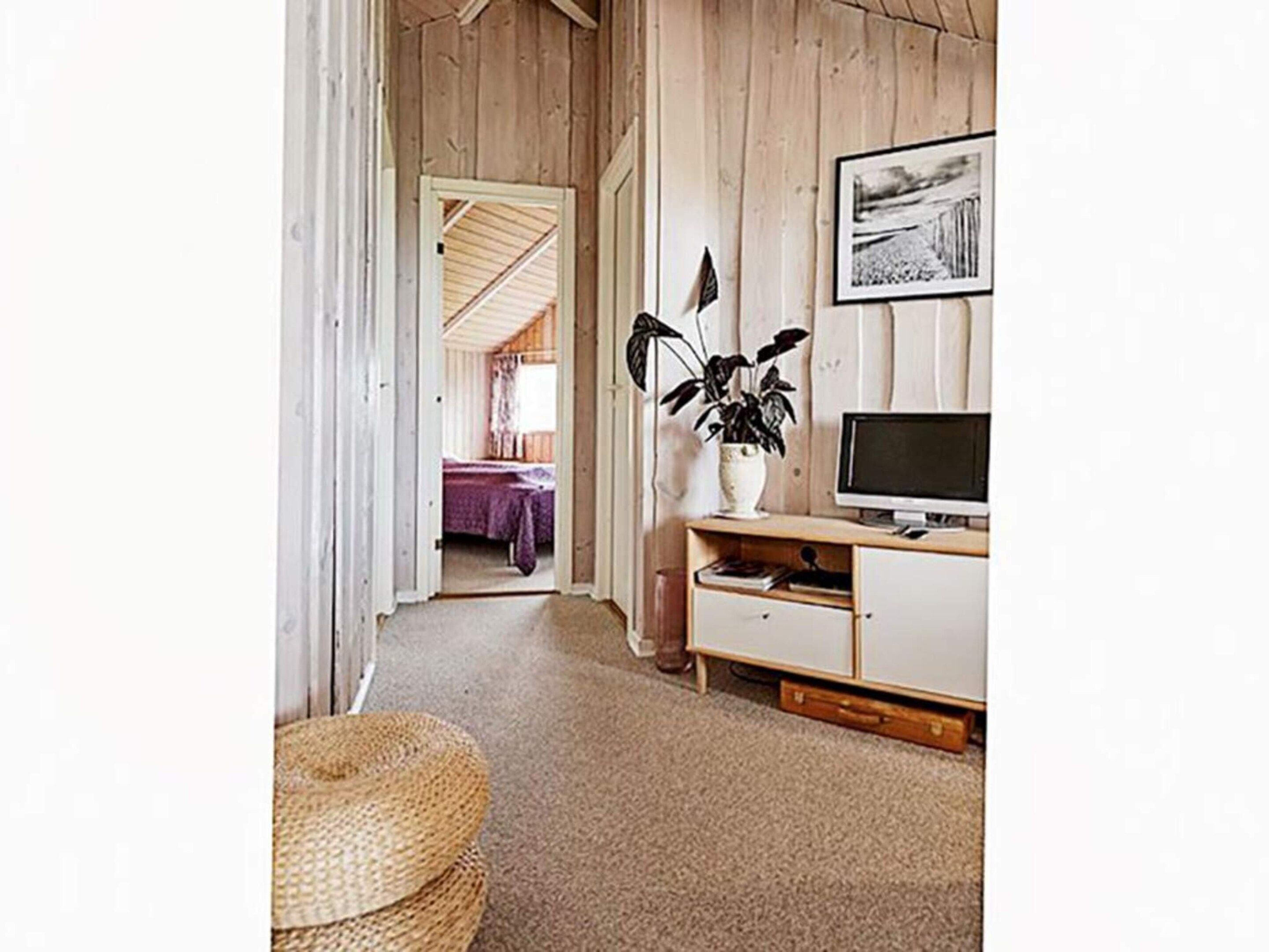 Ein Zimmer mit Holzboden, einem Fernseher auf einem Holztisch, einer Pflanze auf einem weißen Schrank und einem Durchgang zu einem weiteren Raum.