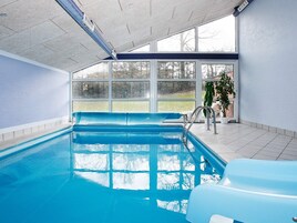 Uma piscina interna com paredes de vidro transparente, piso ladrilhado e vista para o exterior.
