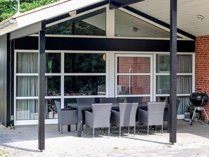 A covered outdoor dining area with a table and chairs, large windows, and a brick wall. - 5 Star Holiday Home in Hadsund (Hadsund)