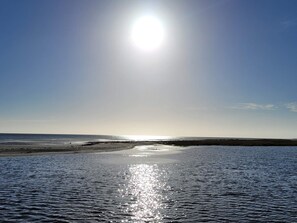 Una playa con agua tranquila que refleja la luz del sol, un cielo despejado y tierra a lo lejos.