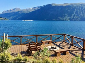 Une terrasse en bois, dotée de bancs et d’un foyer, offre une vue sur un lac, avec des montagnes en toile de fond.