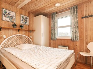 A bedroom with a wooden ceiling, a bed with a white headboard, a window with curtains, and two wall-mounted artworks.
