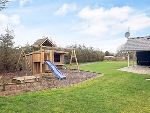 A wooden playhouse with a slide, a swing set, and a covered seating area.