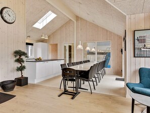 A spacious dining area with a long table and chairs, a clock on the wall, and a view of the city through large windows. - 5 Star Holiday Home in Vejby (Vejby)