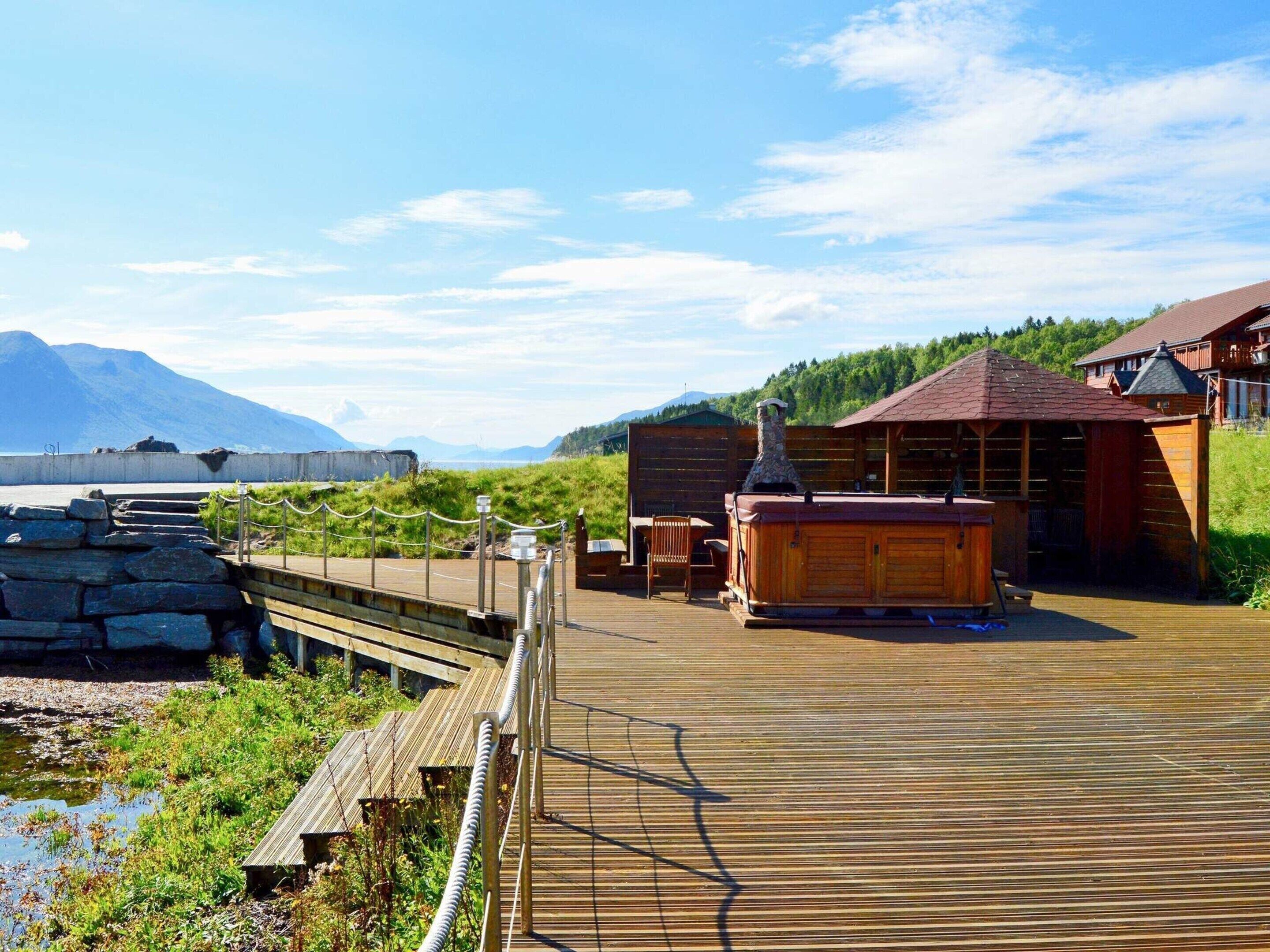 A wooden deck with a hot tub and outdoor seating, overlooking a body of water and mountains.