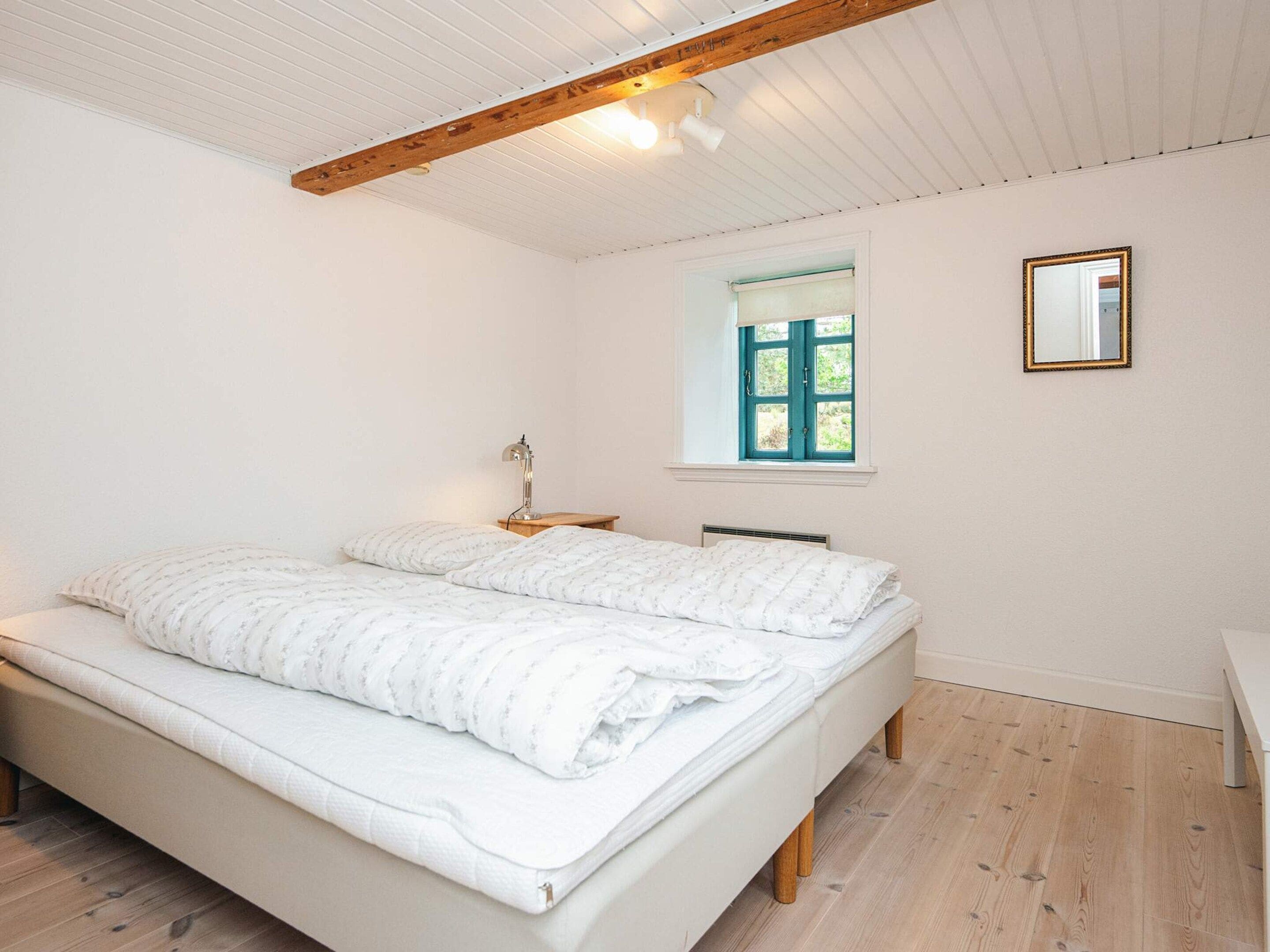 A bedroom with a bed, a bedside table, a lamp, a window, and a mirror.