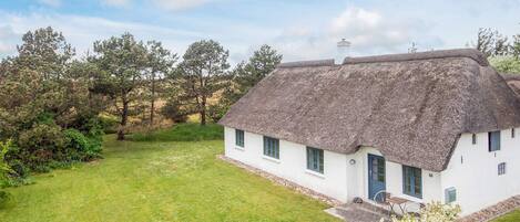 A thatched-roof house with a white exterior, surrounded by a grassy lawn and trees.