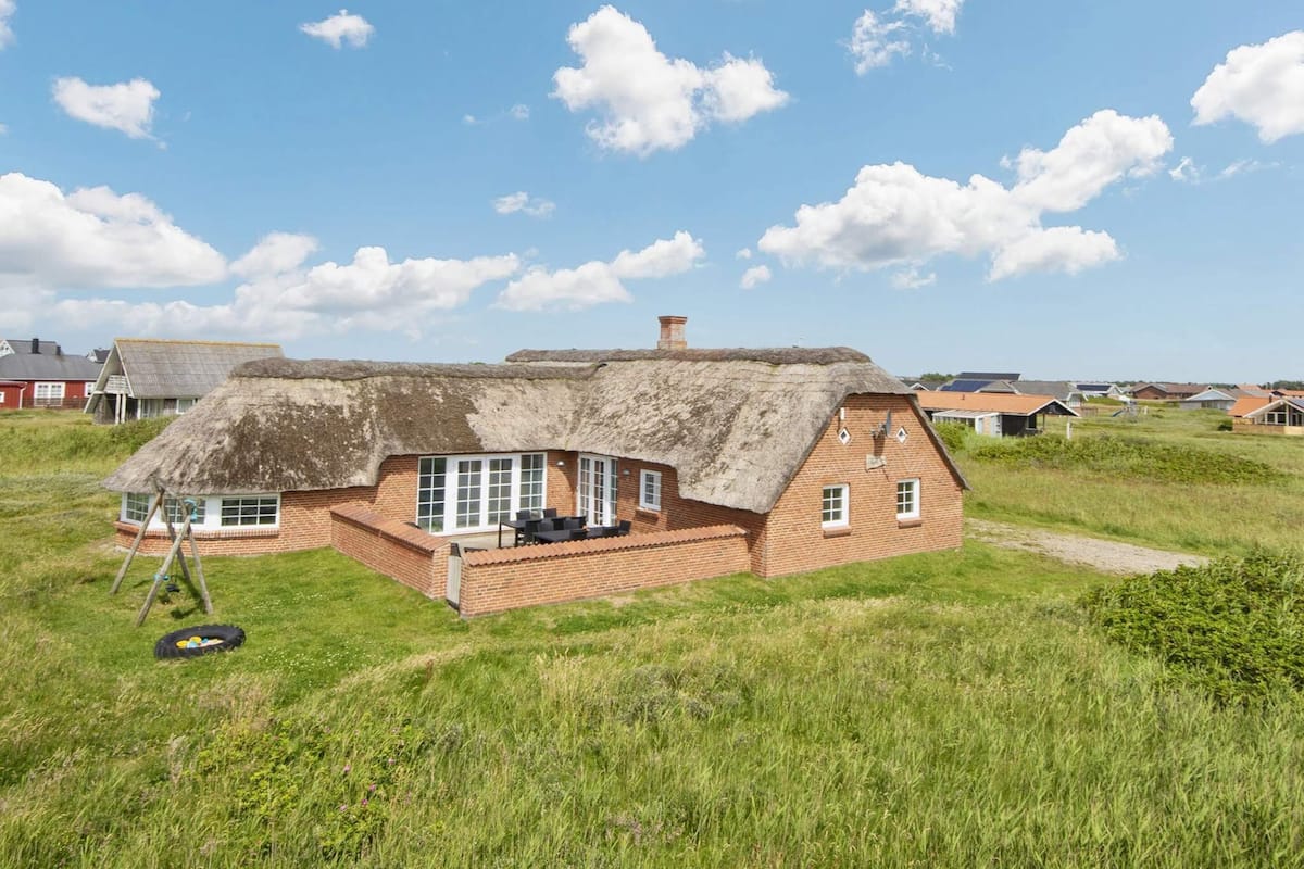 A thatched-roof house with a brick base, surrounded by a grassy field and other houses in the background.