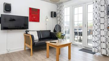 A living room with a black leather sofa, a wooden coffee table, a TV mounted on the wall, and a door leading to an outdoor area.