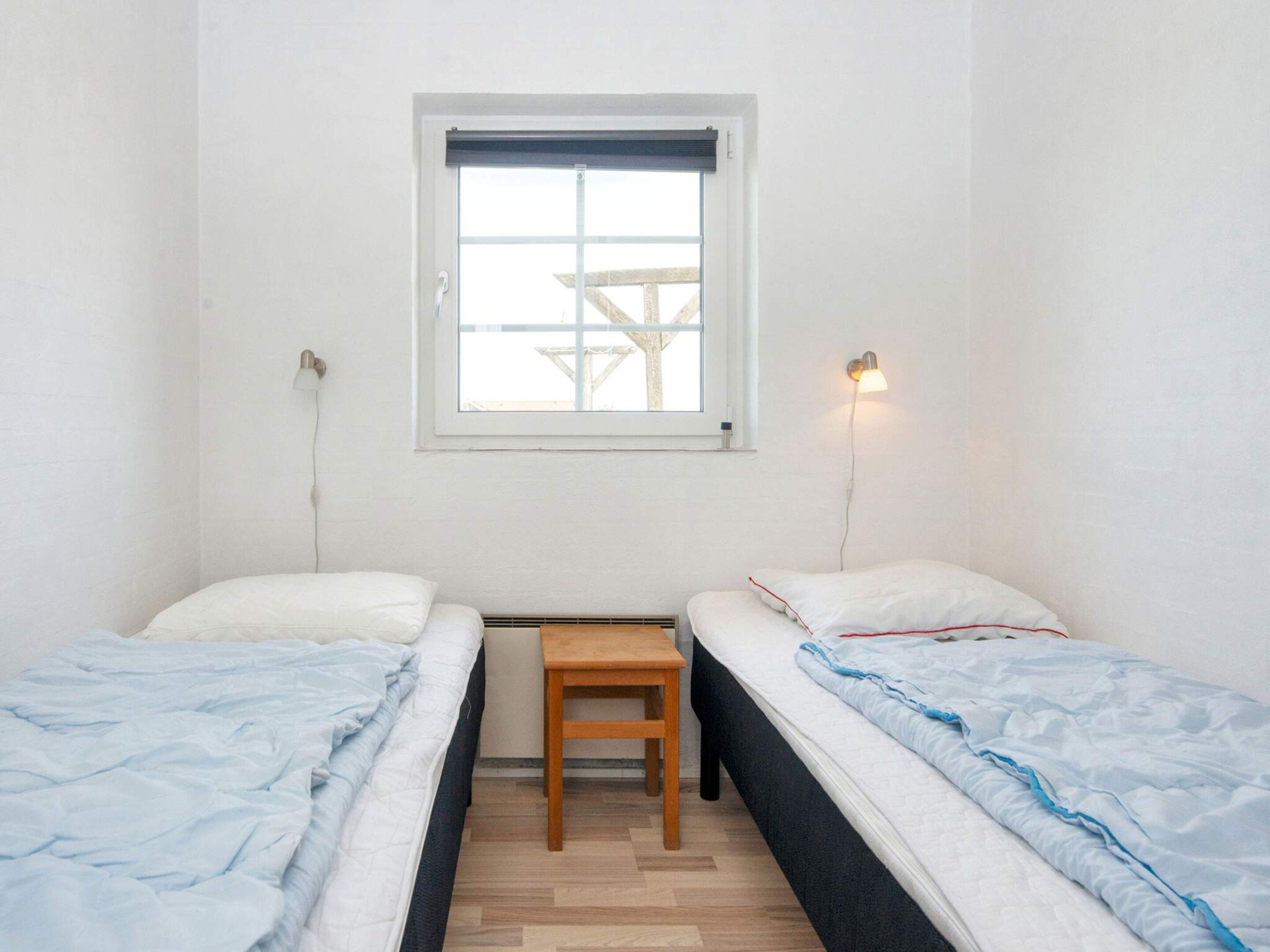 A small, clean room with two single beds, a small wooden table, and a window with blinds.