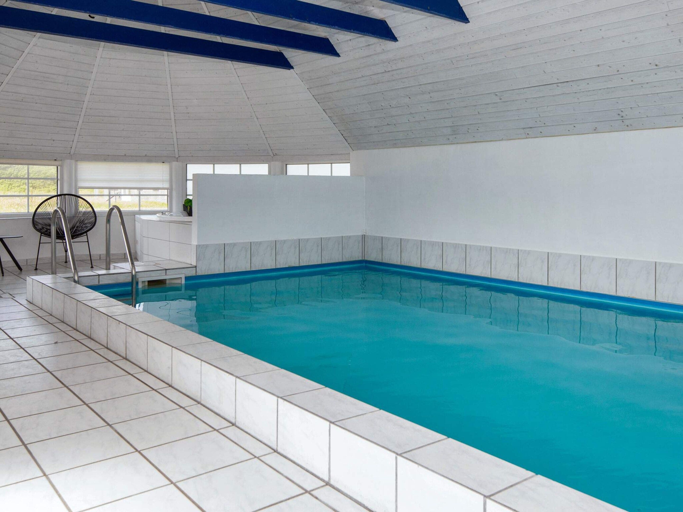 An indoor swimming pool with a tiled floor and walls, a metal ladder, and a chair.