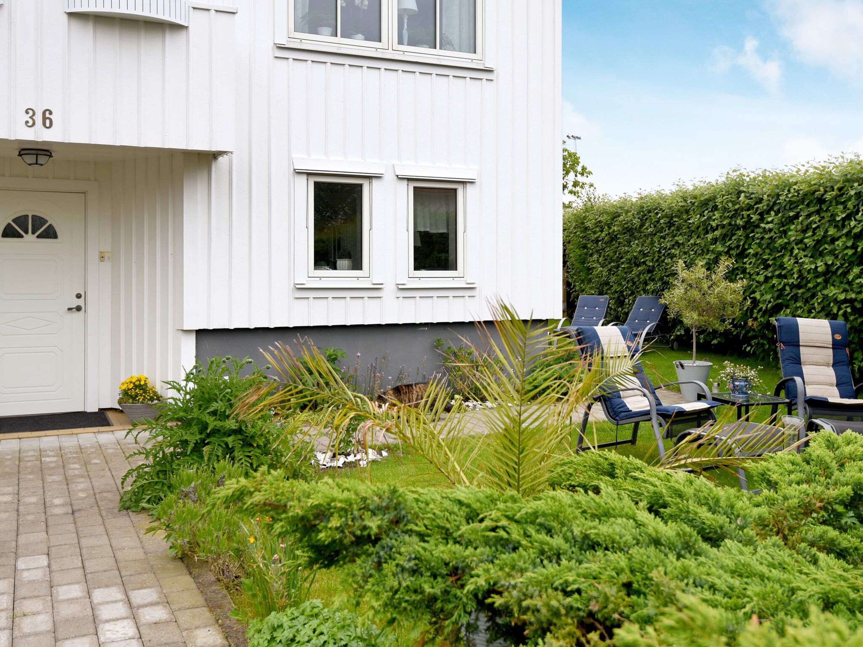 Una casa blanca de dos pisos con el número 36 en la puerta, un jardín con diversas plantas y un área de estar exterior con sillas y una mesa.