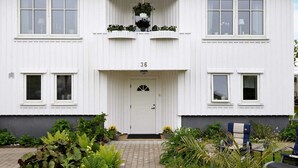A white two-story building with a number 36 on the front door, a small balcony with plants, and a garden with various plants and a bench. - 3 Person Holiday Home in Lysekil (Lysekil)