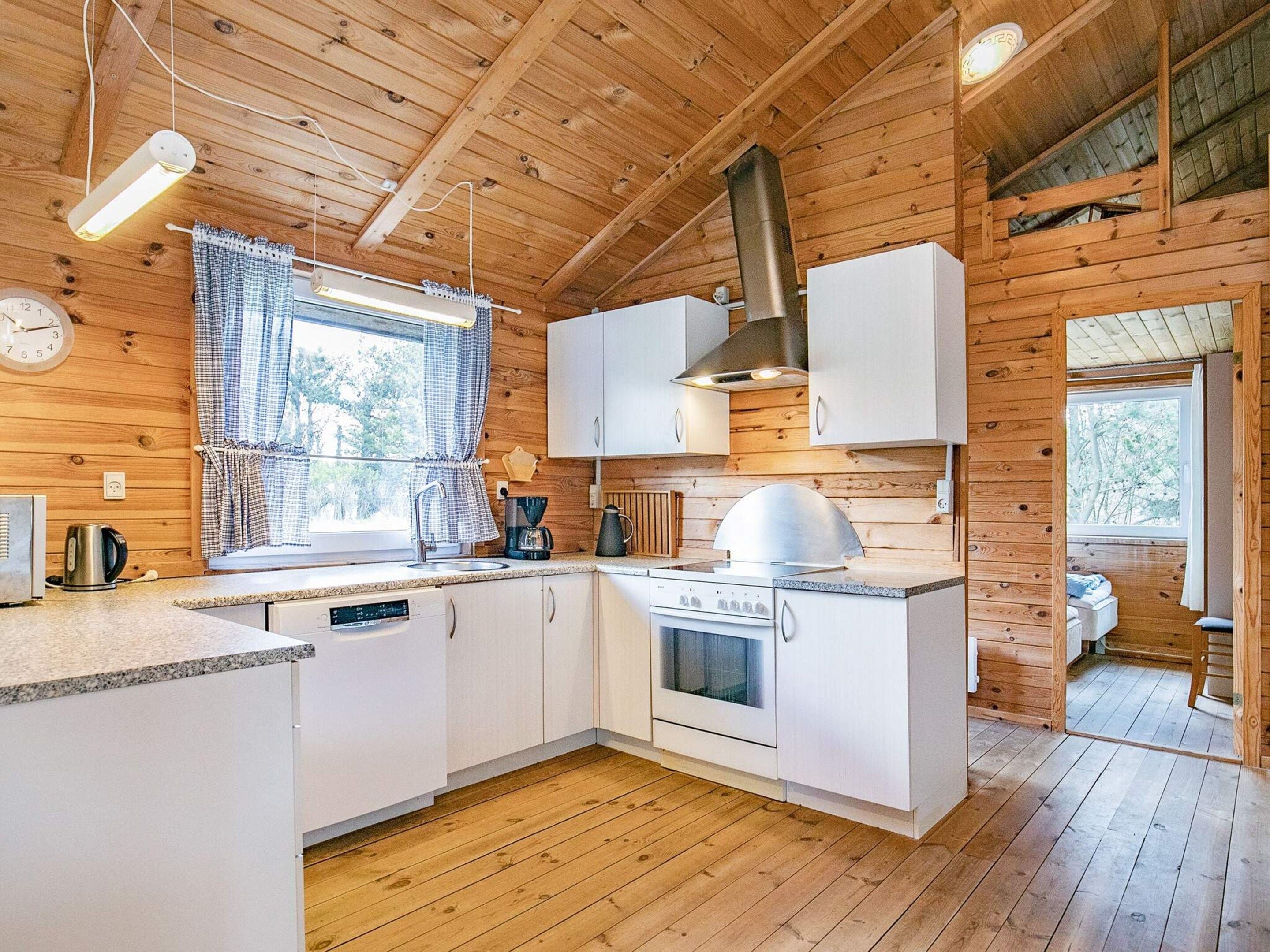 L’intérieur d’un chalet en bois comprend un coin cuisine avec des meubles blancs, un poêle et une fenêtre agrémentée de rideaux.