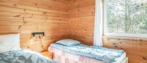 A cozy cabin room with wooden walls, a large window, a bed with blue bedding, and a bedside table with a lamp.
