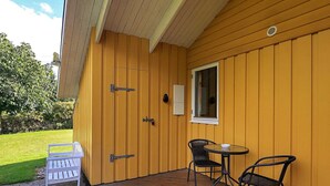 Eine Holzterrasse mit einer weißen Bank und zwei Stühlen, einem Fenster und einer Tür mit Metallscharnieren.