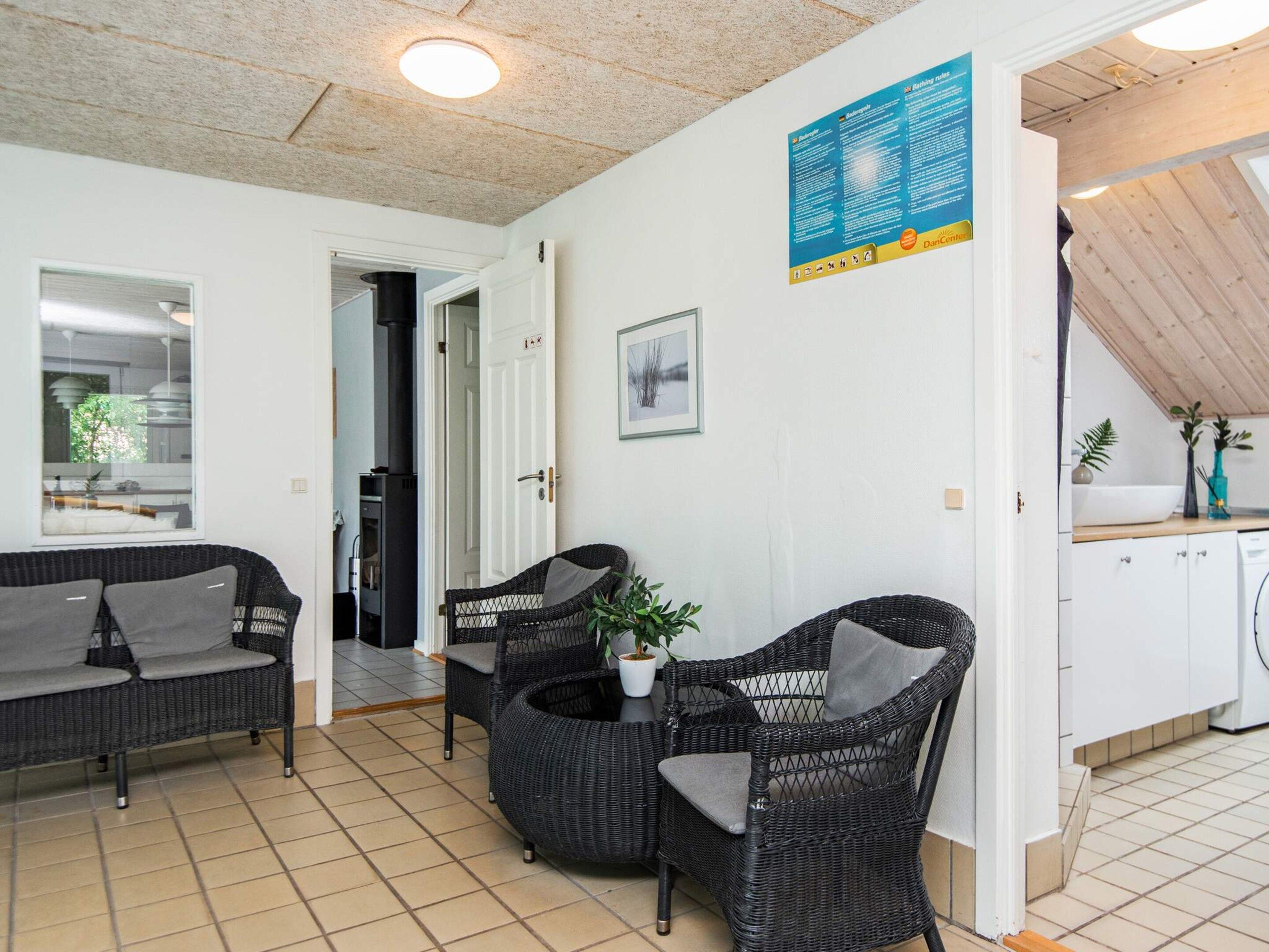 A room with a tiled floor, a sofa, two wicker chairs, a potted plant, and a kitchen area with cabinets and a sink.