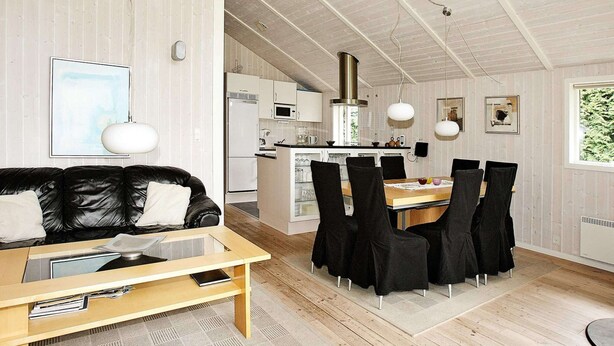 A modern living room with a black leather sofa, a wooden coffee table, and a dining area with black chairs. A kitchen with white appliances is visible in the background. - Cozy Holiday Home in Hadsund near Family Friendly Beach (Hadsund)