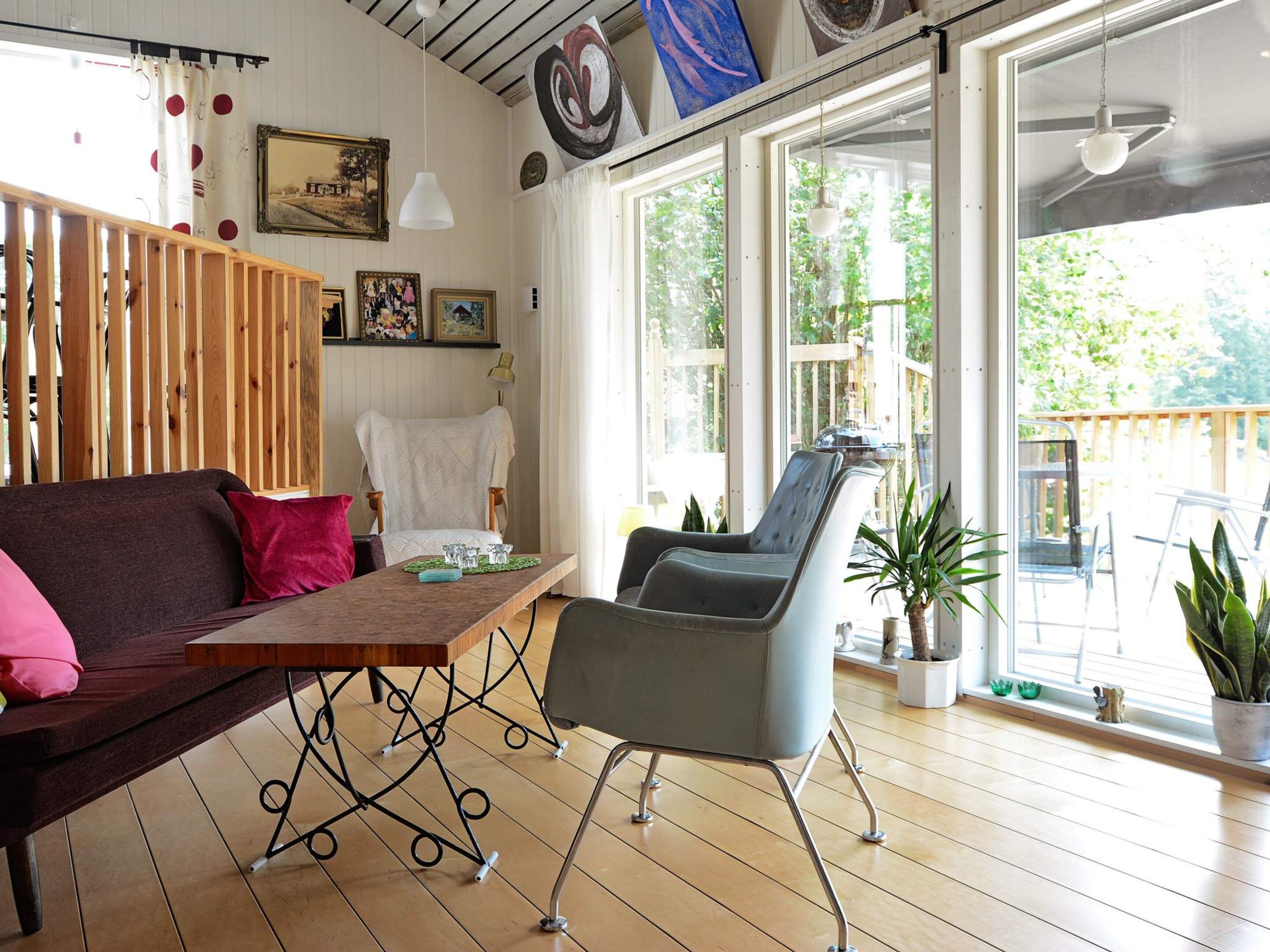 Una habitación con suelo de madera, un sofá, una mesa de comedor y una silla. Hay ventanas con cortinas, un cuadro y una planta.