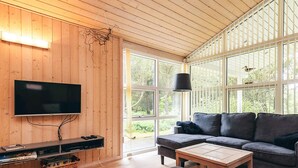 A modern living room with a wooden ceiling, a flat-screen TV, a sofa, a coffee table, and large windows offering a view of greenery.