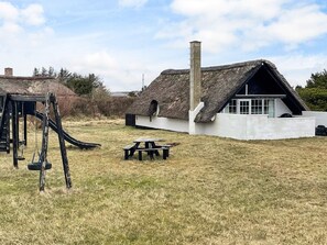 A thatched-roof house with a chimney, a swing set, and a picnic table in a grassy yard. - 6 Person Holiday Home in Ringkobing (Ringkobing)