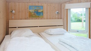 A room with wooden walls, a bed with white bedding, a painting on the wall, and a window with a view of greenery.