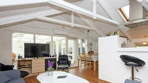 A spacious living area with a large TV, comfortable seating, a dining table, and a kitchen area with a modern design.