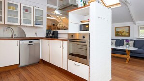 A modern kitchen with white cabinetry, a dishwasher, an oven, and a microwave. There is a dining area with a blue sofa and a table.