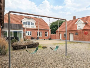 A playground with swings and a gravel area in front of a brick building with a red-tiled roof.