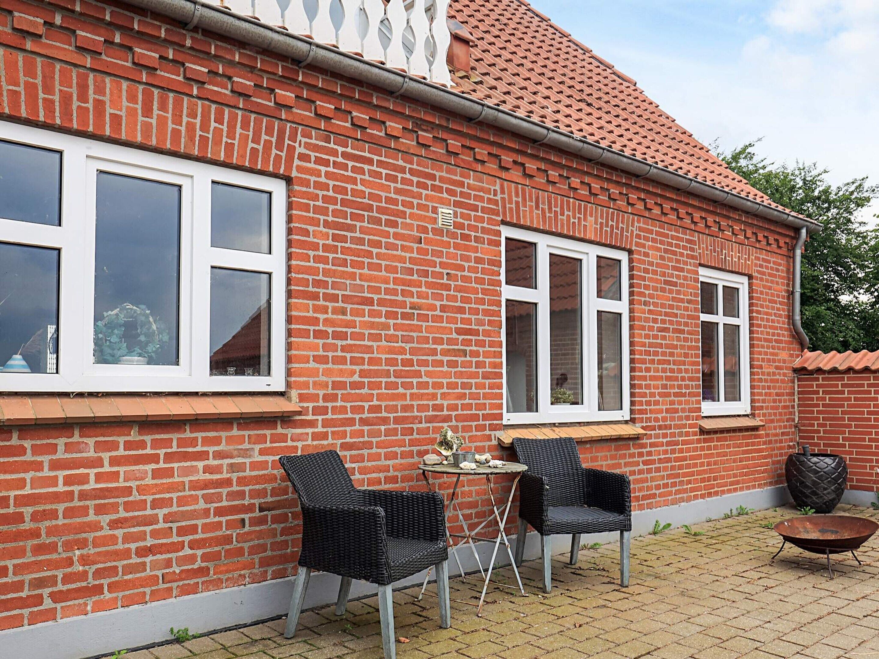 A brick house with two windows, a small table, two chairs, and a wheelbarrow.