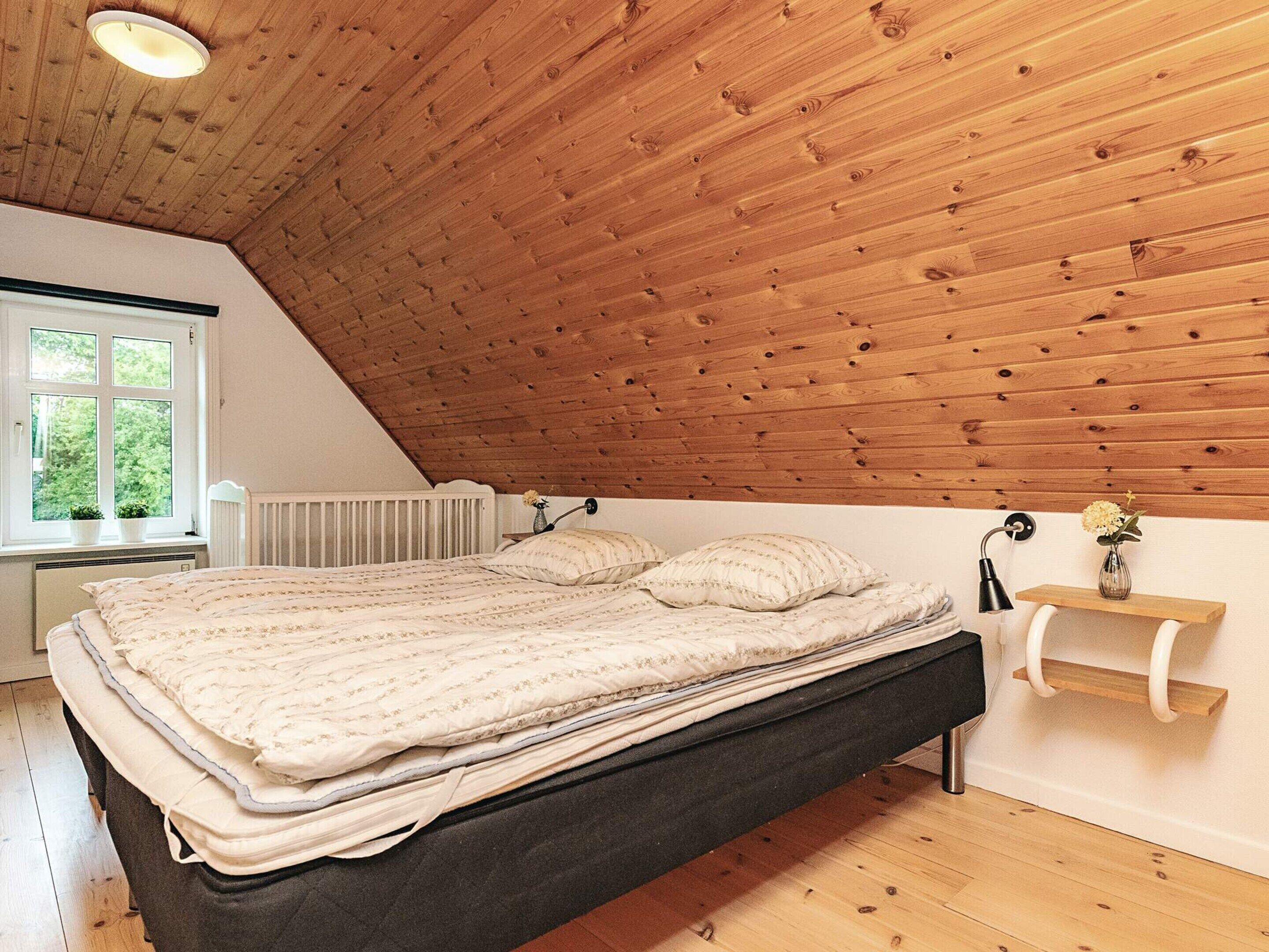 A bedroom with a sloped ceiling, a bed with a dark mattress, a bedside table with a lamp, and a window with a view of greenery.