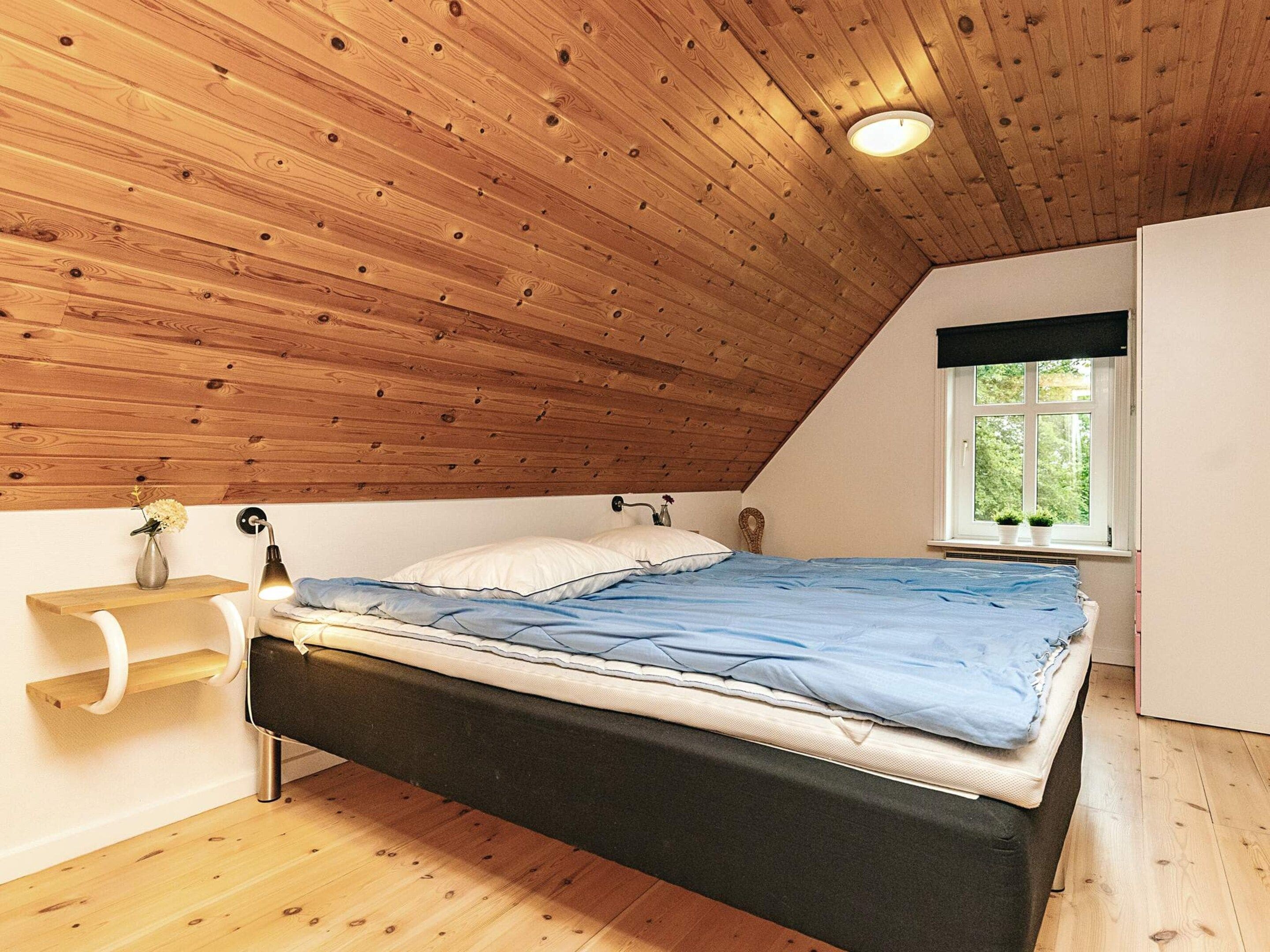 A bedroom with a sloped ceiling, a bed with a blue mattress, a bedside lamp, and a window with a view of greenery.