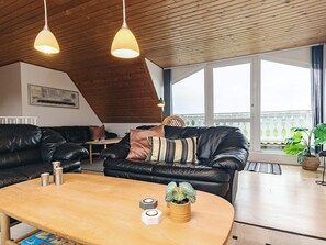 A modern living room with a wooden ceiling, leather sofas, a wooden coffee table, and large windows offering a view.