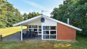 A small, single-story building with a brick exterior and a white roof. It features a covered patio with outdoor seating and a satellite dish on the roof. - 8 Person Holiday Home in Oksbol (Oksbol)