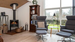 A modern living room with a wood-burning stove, two grey armchairs, a bookshelf, and a large window with a view of greenery. - 8 Person Holiday Home in Oksbol (Oksbol)