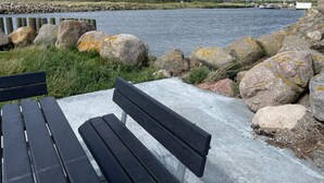 Un banc situé au bord d’un plan d’eau, avec une rive rocheuse et une vue lointaine sur un bâtiment.