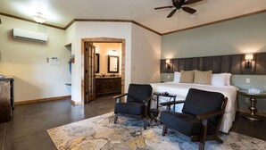 Une chambre à coucher comprenant un lit, deux fauteuils, une petite table, un ventilateur de plafond et une porte en bois donnant sur une salle de bain.