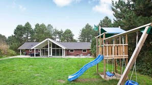 A backyard with a wooden play structure, including a slide and swing set, and a house with a covered patio. - Spacious Holiday Home in Rødby near Beach (Rodby)