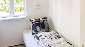 Pieni huone, jossa on yksi sänky, ikkuna, seinään kiinnitetty valaisin ja sanomalehtikuviollinen tyyny.