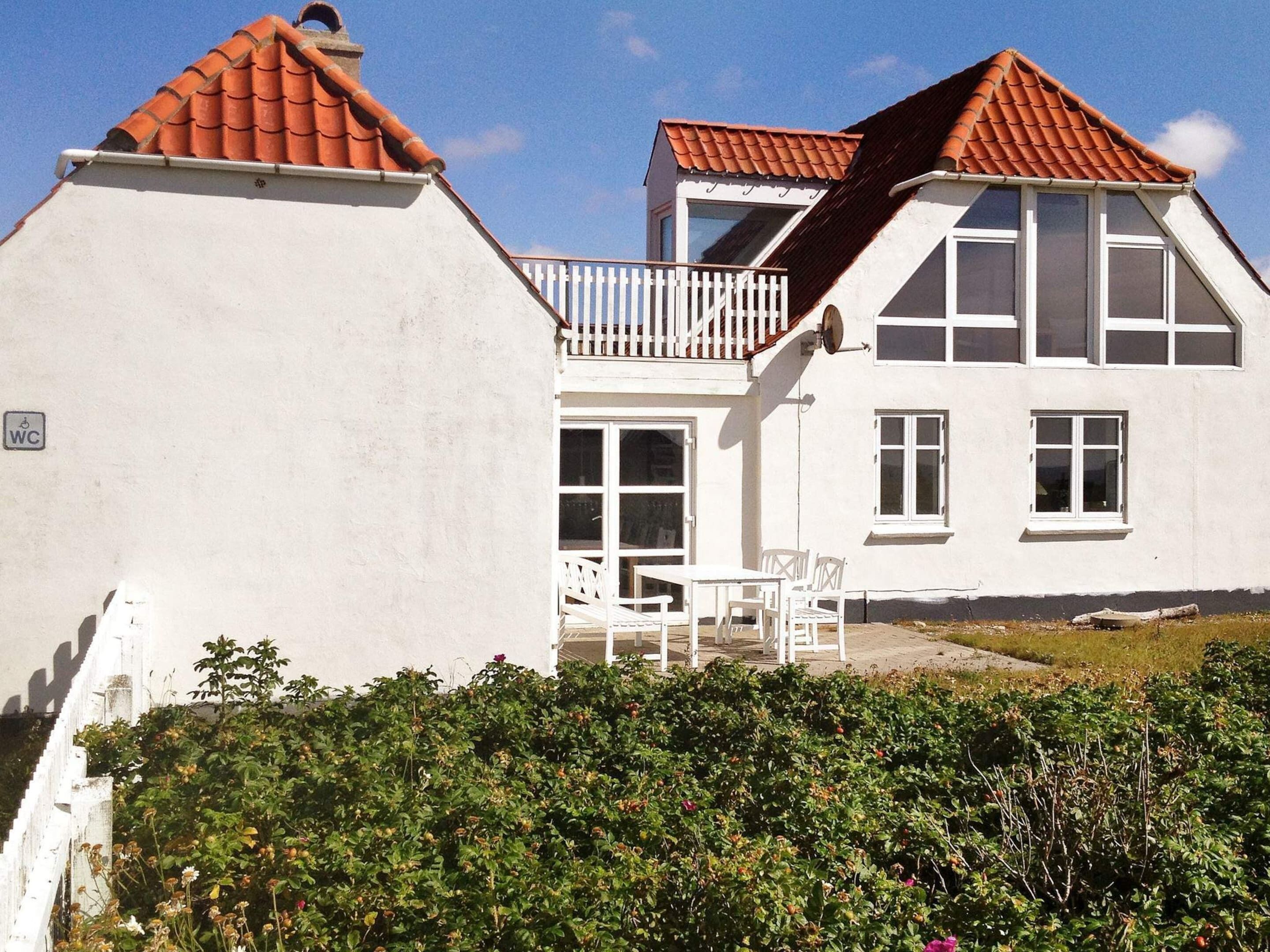 A two-story white building with a red-tiled roof, a balcony with white railings, and a small garden area with bushes and flowers.