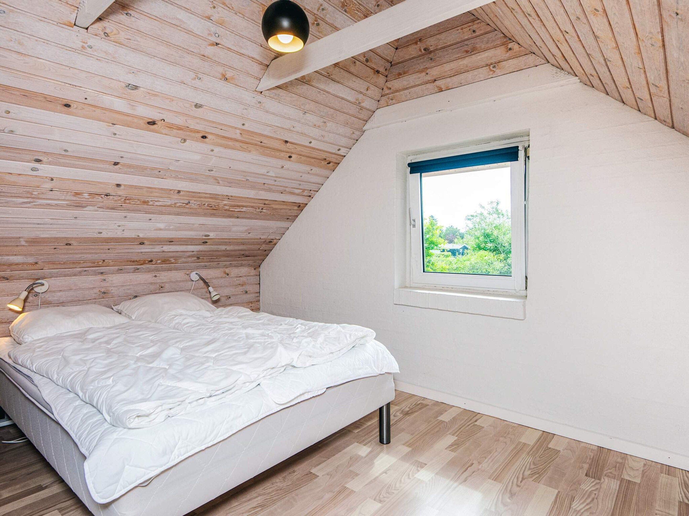 A bedroom with a sloped ceiling, a bed with white bedding, a small window with a view of greenery, and a wall-mounted light fixture.