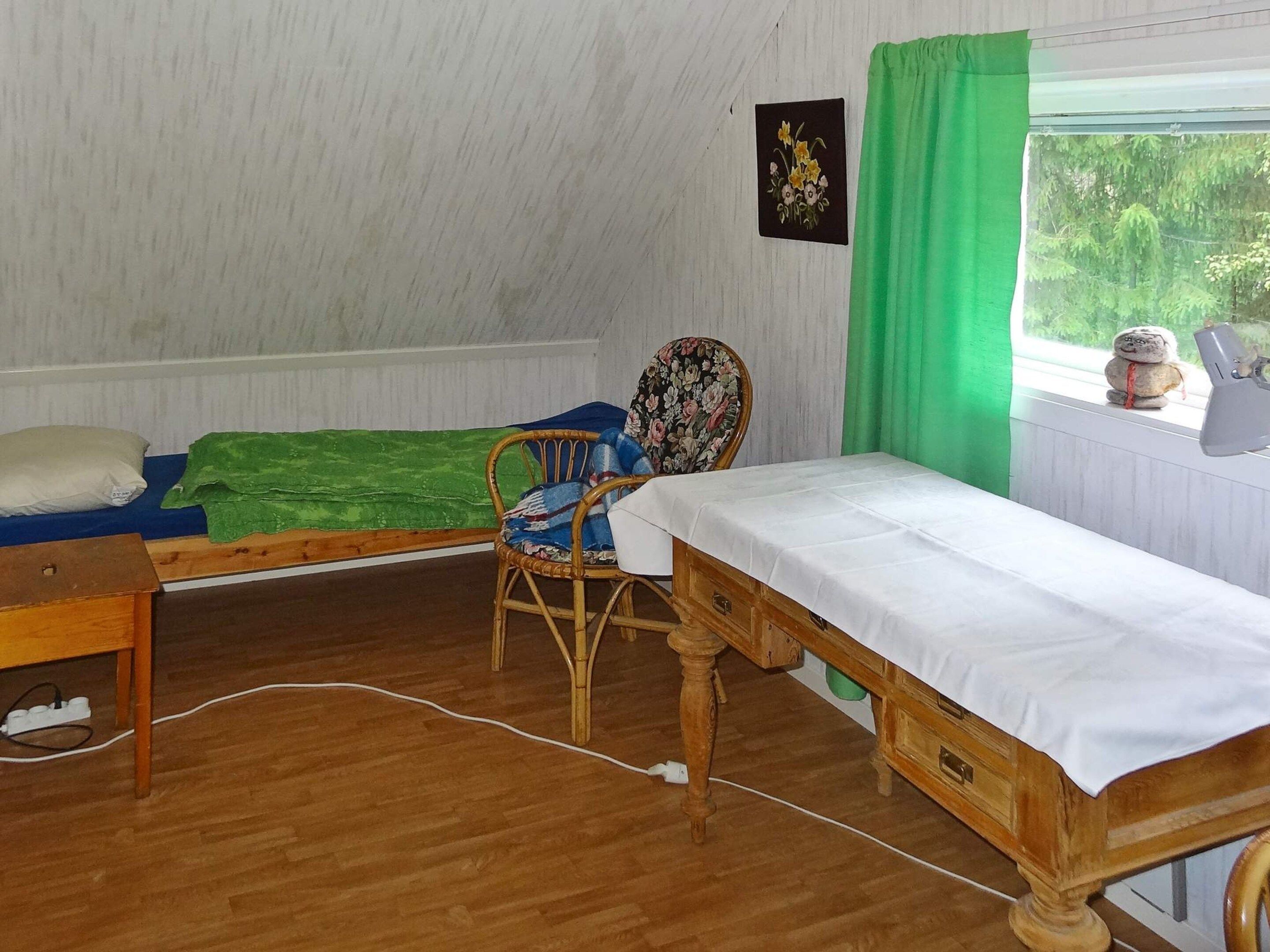 A room with a wooden floor, a bed with a white sheet, a chair with a floral pattern, a small table, and a window with a green curtain.