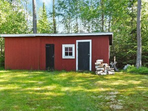 A red wooden shed with a black door and a small window, surrounded by a grassy area and trees. - 5 Person Holiday Home in Logdea (Lögdeå)