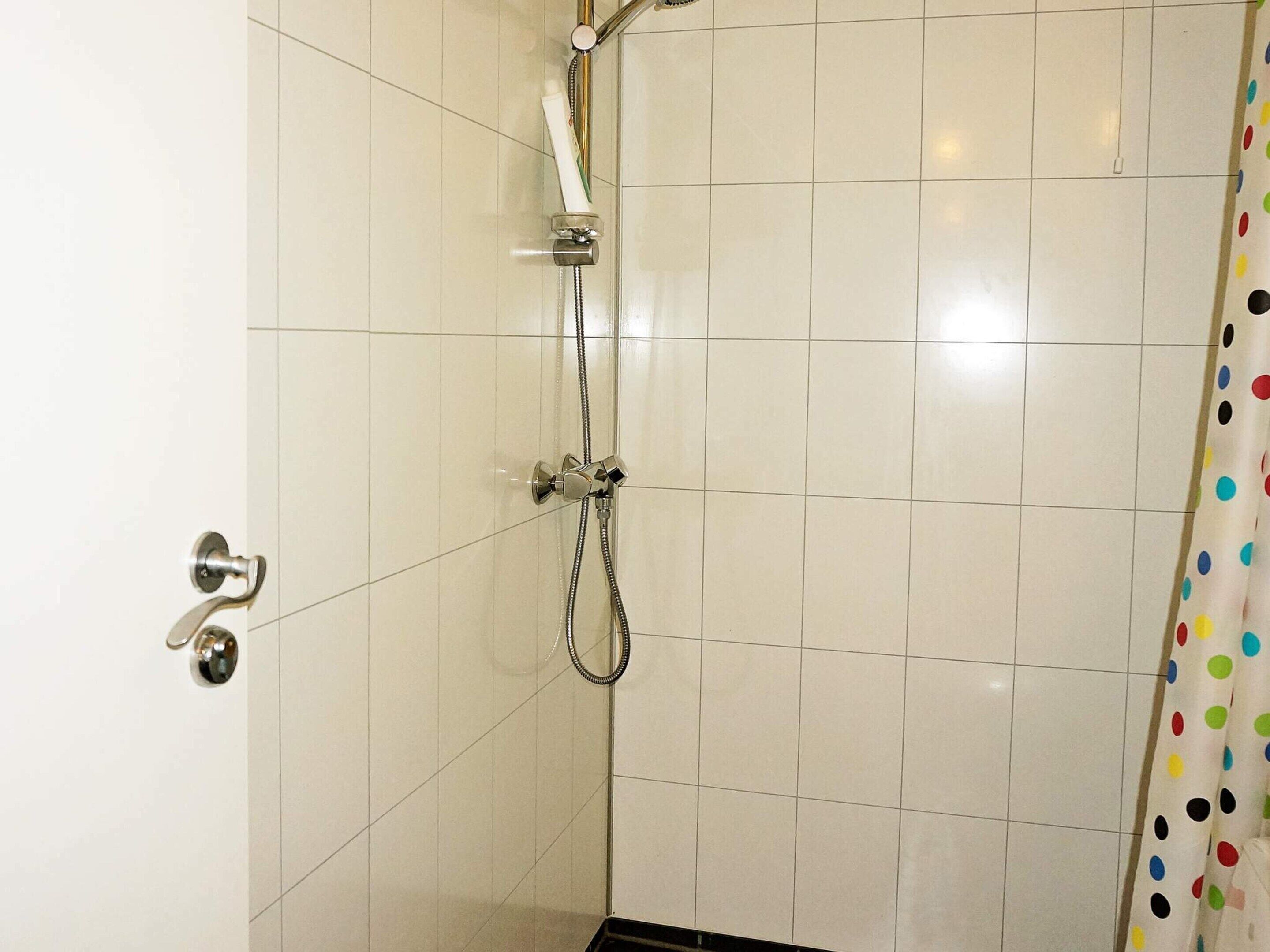 A shower with a handheld showerhead and a wall-mounted faucet, tiled walls, and a door with a silver handle.