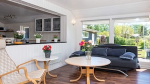 Ein modernes Wohnzimmer mit Holzboden, einem runden Tisch, einer Couch und einem Sessel. Durch einen offenen Durchgang ist eine Küchenzeile mit Schränken und einer Arbeitsplatte erkennbar.