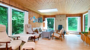 En rummelig stue med trægulv, store vinduer og et hyggeligt opholdsområde med flere stole og en sofa.