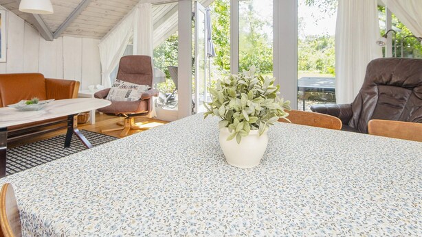 A cozy room with a wooden ceiling, a round table with a floral tablecloth, two brown leather chairs, a white armchair with a newspaper cushion, and a potted plant. - 4 Person Holiday Home in Oksbol (Oksbol)